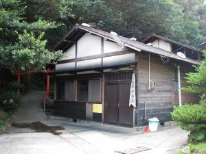 左奥には赤い鳥居が見え、緑豊かな森の中に木造の古い日本家屋が建っている湯向温泉の外観写真