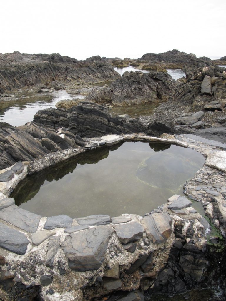 岩場に囲まれた海辺に円形の石組みの潮だまりがあり、静かな水面に周囲の黒い岩が映り込んでいる写真