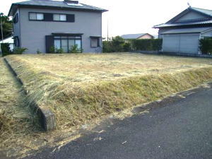 前面は刈り取られた雑草が乾燥した状態の、平らな土地が広がり、奥には二階建ての住宅と平屋の建物が見える、住宅地の空き地を捉えた写真