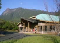 青い空の下、緑に囲まれた山を背景に、木造とモダンな屋根を組み合わせたデザインの建物が建ち、手前には芝生と舗装された進入路が写っている、ぽん・たん館の外観写真