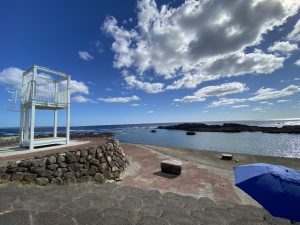 青い空に白い雲が浮かび、海岸沿いの広場に白い監視台と岩を積んだ擁壁、そして青いパラソルの一部が写っている、岩場の海の風景写真