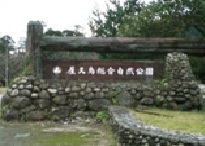 「屋久島総合自然公園」と書かれた木製の看板が、苔むした自然石の台座に設置されており、その左と上には大きな丸太が門のように組まれ、背後には鬱蒼とした木々が見える写真