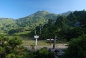 明るい早朝の日差しを浴びて青々と茂る植生が広がる広場があり、そこには木の道標と白い案内板が立っていて、遠景には緑の森に覆われた山がそびえ立つ、登山口付近の景色を捉えた写真