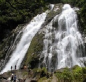 複数の段と岩肌を広範囲にわたって流れ落ちる水しぶきが雄々しい大川の滝の麓に、数名の人が立っている自然の風景写真