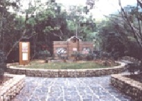 石垣と舗装されたタイルで囲まれた円形の芝生広場の中央に、シンプルな茶色の建物と情報掲示板が建ち、周囲を木々に囲まれている公園内の風景写真