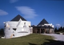 コンクリートの円形と台形を組み合わせたような複雑な構造を持つ現代的な建物が、晴れた青空の下、緑の芝生の上に建ち、その建物の上部には黒いピラミッド型の屋根が二つ見える写真