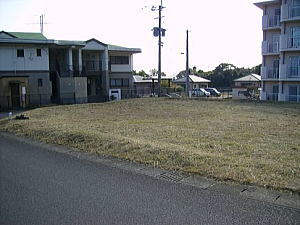 アスファルトの道路の脇に、雑草がまばらに生えた茶色の土の空き地が広がり、その奥には緑色の屋根の住宅や白壁の4階建ての集合住宅が建っている分譲地の写真