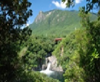 緑豊かな木々の葉の間から、険しい山を背景に、岩場を流れ落ちるトローキの滝とその下の水面が見える、鮮やかな自然の風景写真