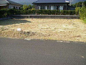 整地された草地の空き地が、手前にアスファルトの道路、奥にはコンクリートの低い塀を隔てて黒っぽい日本家屋が写り、両脇には木々の葉が見える分譲地の写真