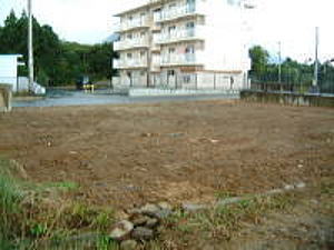 草刈りがされた後の平坦な空き地で、細い道路を挟んだ奥にはベージュ色の4階建ての集合住宅が建っている分譲地の写真