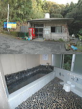 山々の緑を背景に、コンクリート打ちっぱなしの外観を持つシンプルな平屋の建物の写真と、その建物の内部にある黒い玉石が敷き詰められた床と石を縁取った浴槽がある内湯を写した2枚の写真が上下に配置された写真