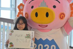 眼鏡をかけた女の子が手書きで「きばれかごしま」と書かれたスケッチブックを掲げ、ピンク色の大きな豚のキャラクターの着ぐるみと並んで、写っている写真