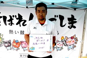 テントの下で白いポロシャツを着た男性が、手書きで「みんながんばって！！応援しています いちご」と書かれた白いボードを持って正面を向いて立っている写真