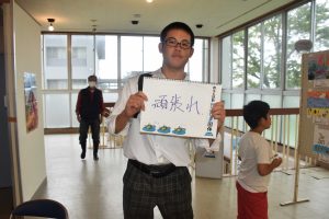 室内のロビーの一角で坊主頭の男の子が手書きで「頑張れ」と書いたスケッチブックを持って立っている写真
