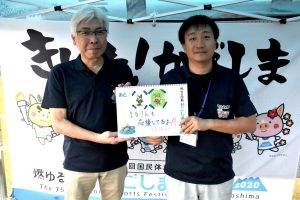 テントの下で黒っぽいポロシャツを着た二人の男性が、左側の男性は白髪でメガネ、右側の男性は黒髪でネームプレートを下げ、緑のキャラクターのイラストと「まうりんも応援してますよ！！」と書かれたボードを中央で一緒に持っている写真