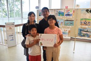 屋内ギャラリーで、父親、母親、2人の子供の4人家族がカメラに向かって並び、子供たちが「がんばれ!」と手書きしたスケッチブックを抱えている写真
