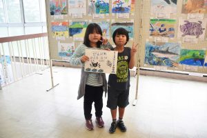 屋内ギャラリーで、2人の子供たちが「がんばれ鹿児島国体」と手書きしたスケッチブックを抱えてピースサインをしている写真