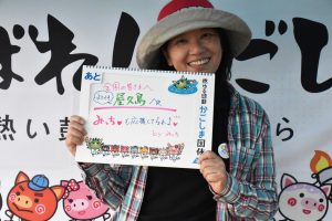 広いつばの帽子を被り、チェック柄のシャツを着た笑顔の女性が、「全国の皆さんへ 燃ゆる感動かごしま国体 in 屋久島へ みちゅも応援してますよ by みちゅ」と書かれたボードを持って立っている写真
