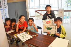 施設内で、6人の子供たちが机を囲み、それぞれがスケッチブックにイラストを描き、2人の子供が手書きのスケッチブックを抱えて皆でピースサインをしている写真