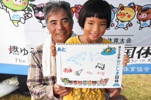 白髪の年配の男性と黄色のシャツを着た女の子が、笑顔で、魚や飛行機の絵と「とべとべー」と書かれたボードを一緒に持って立っている写真