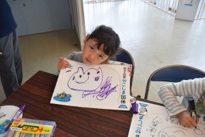 施設内で、子供が机に座り、イラストを手書きしたスケッチブックを抱えている写真