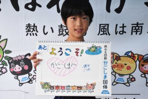 テントの下で前髪を短く切り揃えた小学生くらいの児童が、中央にピンクの大きなハートと「ようこそ！かごしまへ」と書かれたボードを持って立っている写真