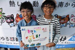 テントの下で、ボーダー柄のシャツを着て眼鏡をかけた女の子、デニムシャツを着て眼鏡をかけた女の子の2人が、「きばれかごしま」と書かれた応援ボードを持って笑顔で並んでいる写真
