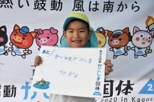 黄色いキャップをかぶり青い服を着た男の子が、満面の笑顔で「がんばれやくしま」自分で書いたボードを胸の前で持ち、立っている写真