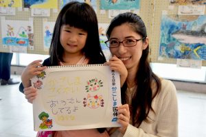 施設内で、眼鏡をかけた母親と子供が手書きのスケッチブックを一緒にかかげている写真