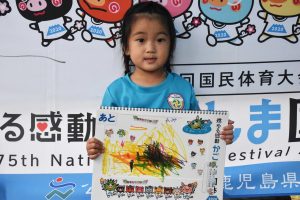 青いTシャツを着た幼い女の子が、自作のカラフルな絵を描いたボードを持って、正面を向いている写真