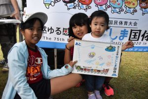 芝生の上で、左の白いキャップをかぶった女の子と、もう1人の女の子で立っている幼い子を支えながら、子供の絵が描かれたスケッチブックを正面に見せている写真