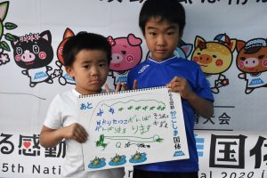 白いTシャツを着た左の幼い男の子と、青い服を着た右の男の子の二人が、カヌーの絵と「ガンバるコトば!!」などの文字が描かれたスケッチブックを正面に見せている写真