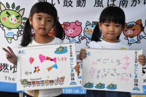 ツインテールにした女の子二人が並んで立ち、左の女の子がハートや星の絵、右の女の子が「かごしまへ ようこそ」などのメッセージが書かれたスケッチブックをそれぞれ正面に向けている写真