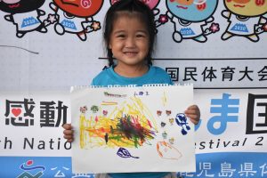 国体の横断幕の前で、青いTシャツを着た笑顔の幼い女の子が、カラフルなクレヨンで描かれた絵を持って立っている写真