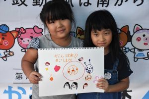 国体の横断幕の前で女の子二人で「がんばれかごしま」と書かれたスケッチブックを持っている写真