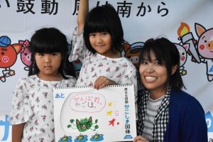 国体の横断幕の前で、お揃いの白いTシャツを着た2人の女の子と、紺色のカーディガンを羽織った女性が、「がんばれかごしま」と手書きした絵を手に笑顔で並んでいる写真
