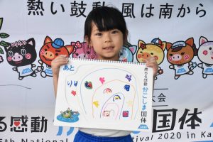 幼い女の子がカラフルな色で描かれた絵を両手で持ち、笑顔で正面を向いている写真