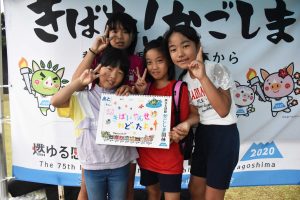 国体の横断幕の前で、笑顔の女の子4人が、「きばいやんせ」と書かれた手書きのカラフルなボードを中央に持ち、ピースサインをして写っている集合写真