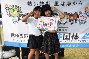 国体の横断幕の前で、女の子二人で手書きの可愛いイラストと文字が描かれたスケッチブックを抱え、満面の笑みでピースサインしている写真