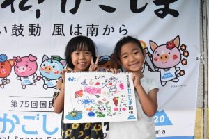 国体の横断幕の前で、女の子二人で手書きの可愛いイラストが描かれたスケッチブックを抱え、笑顔で写っている写真