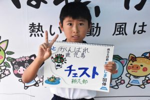 国体の横断幕の前で、男の子が手書きの「がんばれ鹿児島チェスト」と書かれたスケッチブックを抱え、ピースサインをして写っている写真