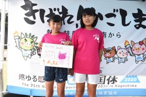 国体の横断幕の前で、お揃いのピンクのTシャツを着た女の子二人で手書きのイラストが描かれたスケッチブックを抱え、写っている写真