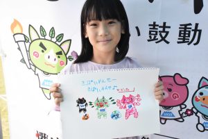 国体の横断幕の前で、女の子が「きばいやんせ」の文字と手書きのイラストが描かれたスケッチブックを抱え、写っている写真