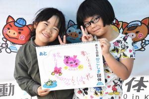 国体の横断幕の前で、眼鏡をかけた女の子とかけていない女の子二人で手書きのイラストと文字が描かれたスケッチブックを抱え、満面の笑みでピースサインしている写真