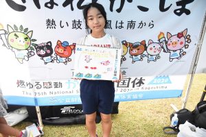 国体の横断幕の前で、女の子が手書きのイラストと文字が描かれたスケッチブックを抱え、立っている写真