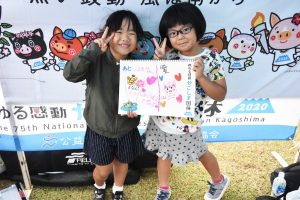 国体の横断幕の前で、眼鏡をかけた女の子とかけていない女の子二人がそれぞれの手書きのイラストと文字が描かれたスケッチブックを抱え、ピースサインしている写真