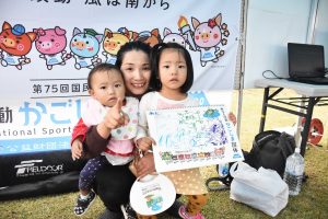 国体の横断幕の前で、母親が二人の子どもを支え、上の子は手書きのイラストが描かれたスケッチブックを抱えている写真
