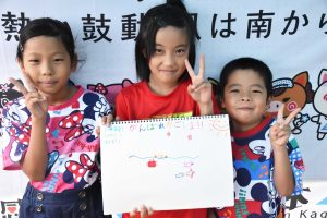 国体の横断幕の前で、3人の子供たちで手書きのイラストが描かれたスケッチブックを掲げ、皆笑顔でピースサインをしている写真