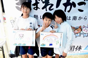 国体の横断幕の前で、白い体操服姿の3人の子供たちが手書きのイラストが描かれたスケッチブックを掲げ、笑顔でピースサインをしている写真
