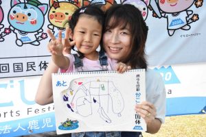 国体の横断幕の前で、手書きのイラストが描かれたスケッチブックを掲げている子どもと抱きかかえている母親が、笑顔でピースサインをしている写真